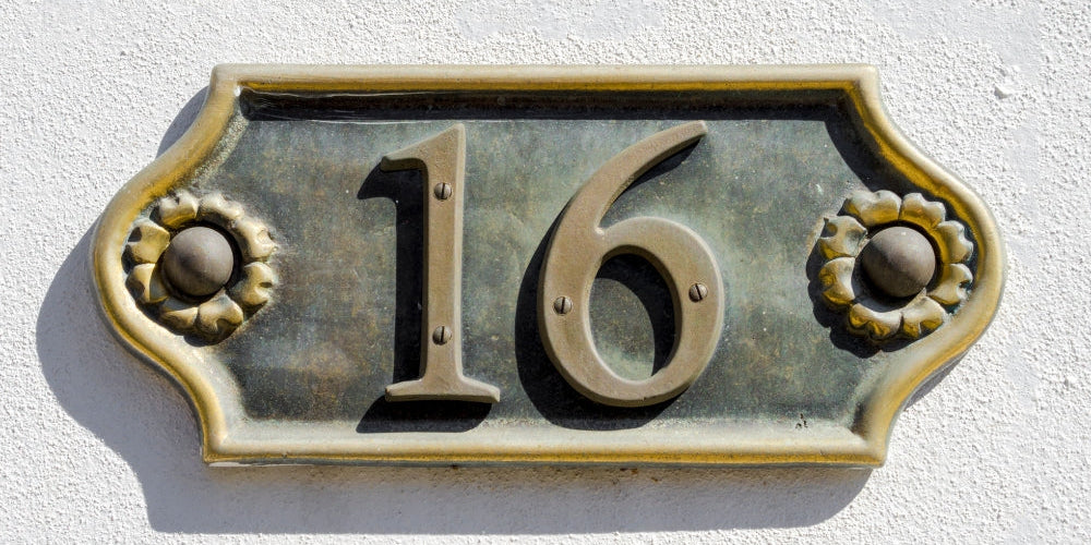 plaque numéro 16 en bronze doré avec fleurs sur mur blanc symbolisant l'heure miroir 16h16 et le courage