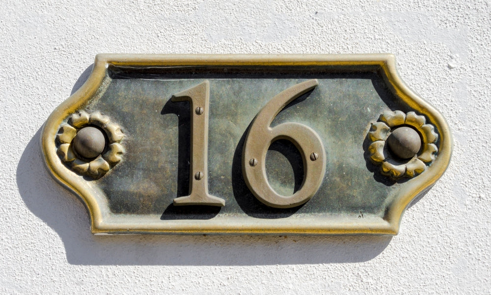 plaque numéro 16 en bronze doré avec fleurs sur mur blanc symbolisant l'heure miroir 16h16 et le courage